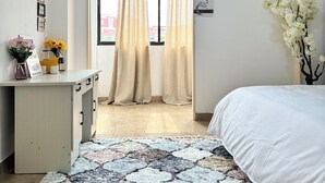 Ein Schlafzimmer mit Bett, Schreibtisch mit Blumenvase, Fenster mit Vorhängen und einer Wand-Klimaanlage.