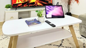 Ein Wohnzimmer mit einem Flachbildfernseher, der einen Kamin zeigt, einem Tablet auf einem weißen Couchtisch, einer Fernbedienung, einem Buch und einer kleinen Pflanze.