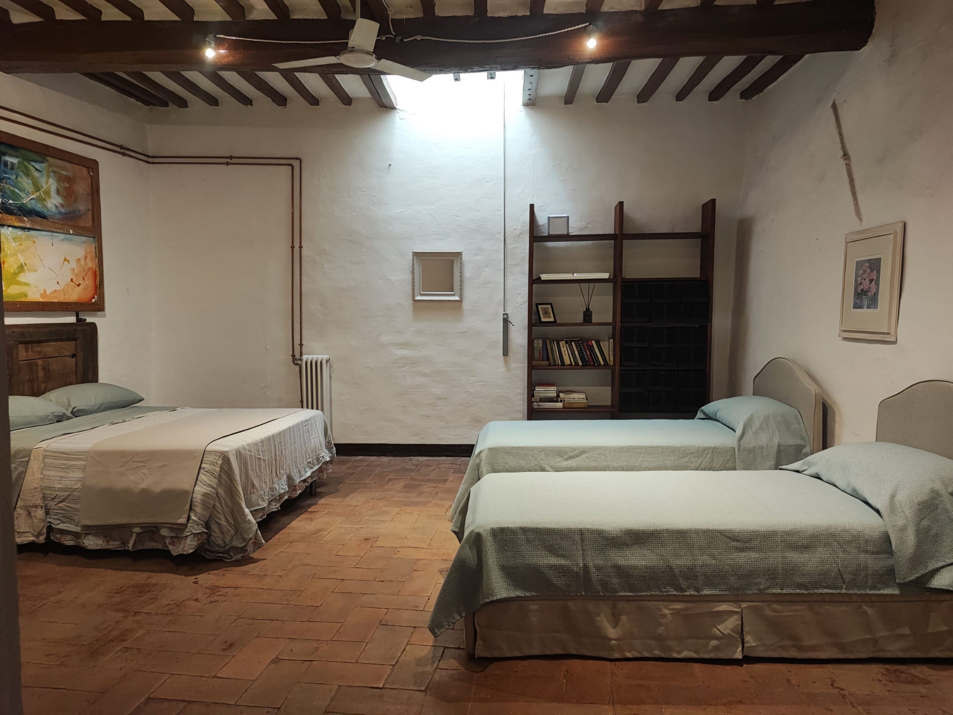 A room with two single beds, a wooden ceiling, a radiator, a bookshelf, and a painting on the wall.
