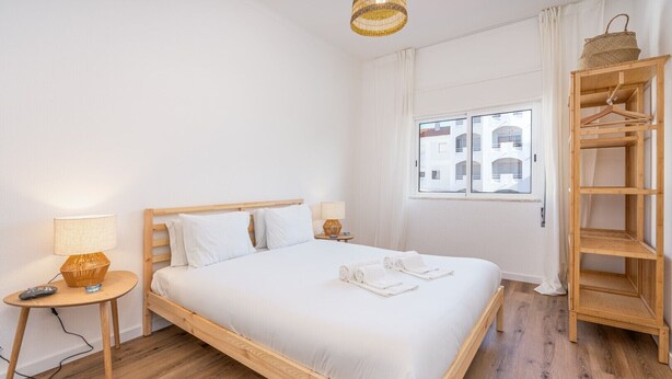 A clean, minimalist hotel room with a wooden bed, bedside lamps, a window with a view, and a wooden shelf with a basket. - #281 Telecomunicações by Home Holidays (Albufeira)
