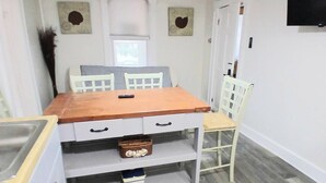 Um quarto pequeno e bem iluminado, com uma mesa de madeira, cadeiras brancas, uma TV fixada na parede e uma janela com persianas.