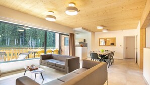 Ein modernes Wohnzimmer mit einer Couch, einem Esstisch und Stühlen. Ein Balkon mit Sitzgelegenheiten bietet Blick auf Bäume.