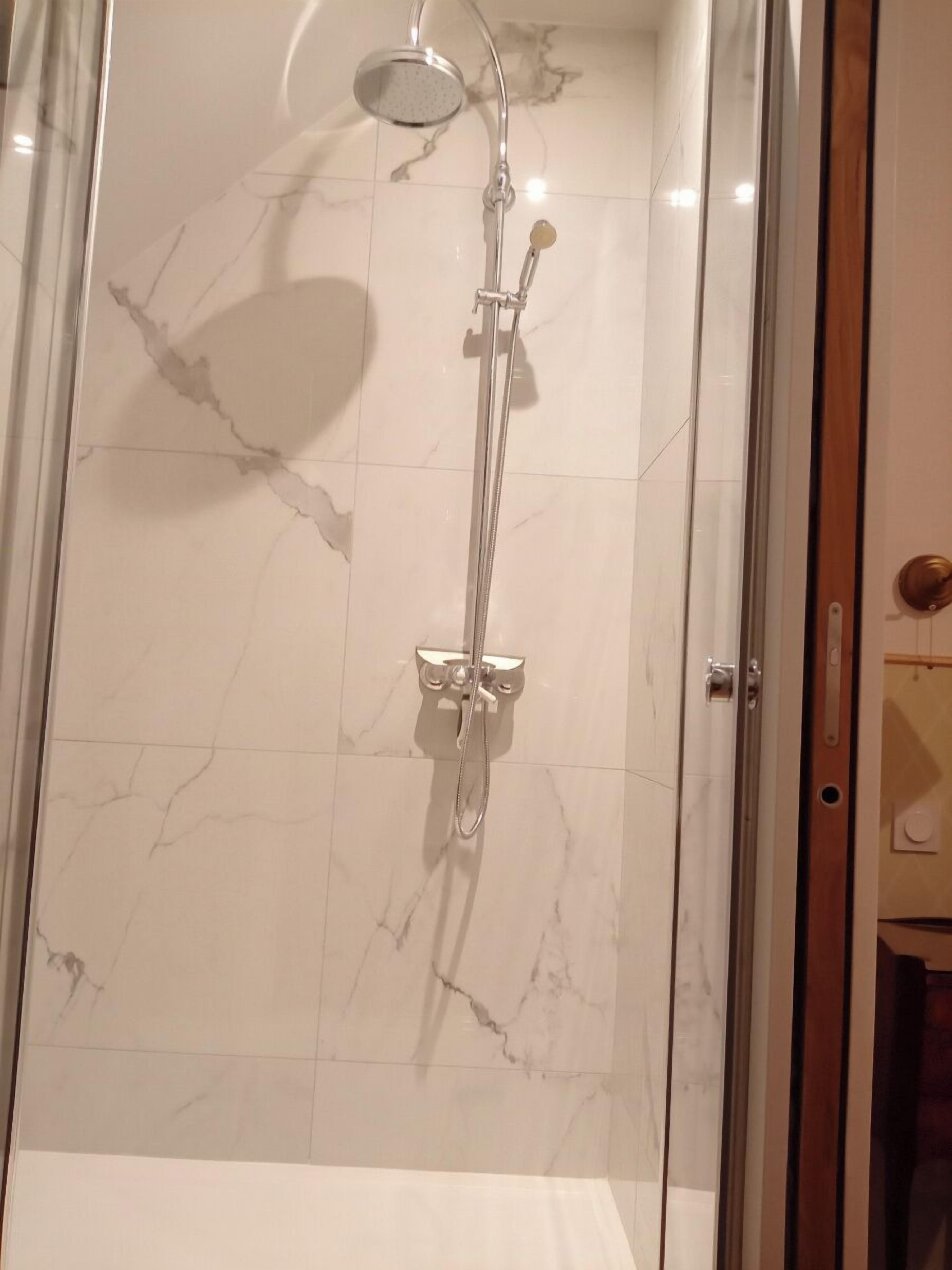 A glass-enclosed shower with a handheld showerhead and a fixed overhead showerhead, set against a marble-tiled wall.