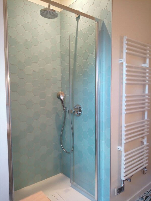 A glass-enclosed shower with a hexagonal tile pattern on the walls, a handheld showerhead, and a wall-mounted towel rack. - à la Mainguère (Brece)