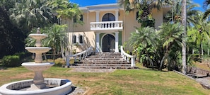 A grand entrance with a stone staircase leading to a balcony, surrounded by lush greenery and a decorative fountain. - Villa Quebrada Estate (Cóbano)