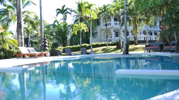 A pool surrounded by palm trees and lounge chairs, with a large building in the background. - Villa Quebrada Estate (Cóbano)