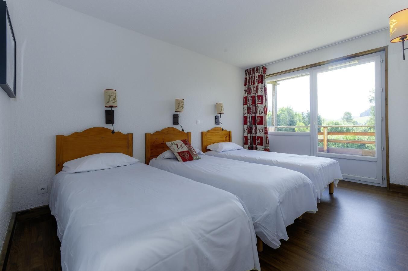 A room with three single beds, each with a wooden headboard and white bedding. There are wall-mounted lamps and a window with a view of greenery.