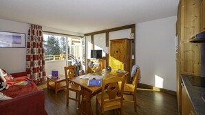 Un salón acogedor con un sofá rojo, una mesa de comedor de madera y sillas. Un televisor está sobre un mueble de madera, y hay una ventana con cortinas.