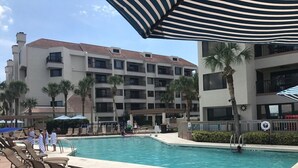 A hotel pool area with multiple lounge chairs, umbrellas, and a building with balconies and palm trees. - Enjoyable 2-bedroom hotel in Hilton Head Island with WiFi (Hilton Head Island)