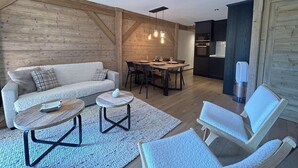 Ein gemütliches Wohnzimmer mit Sofa, Couchtisch und Sesseln, ein Essbereich mit Tisch und Stühlen sowie eine Küchenzeile mit Schränken und Geräten.
