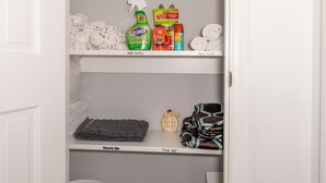 A storage closet with shelves holding rolled towels, cleaning supplies, and a trash bin.