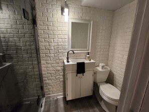 Ein Badezimmer mit weißer Ziegelwand, einem weißen Waschbecken mit Spiegel darüber, einer Toilette und einer Duschkabine.