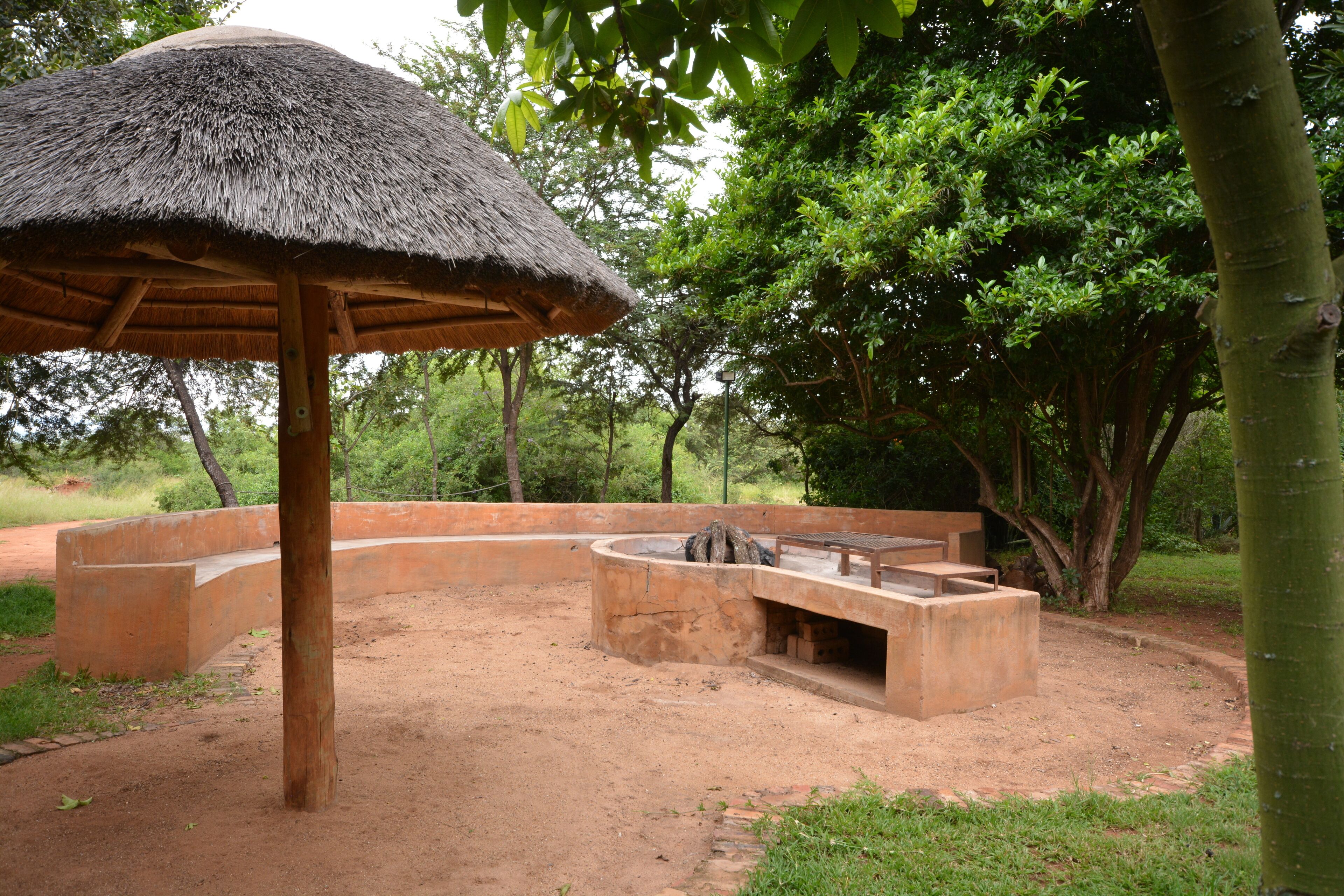 An outdoor area with a thatched-roof structure, a concrete seating area, and a fire pit surrounded by a low wall.