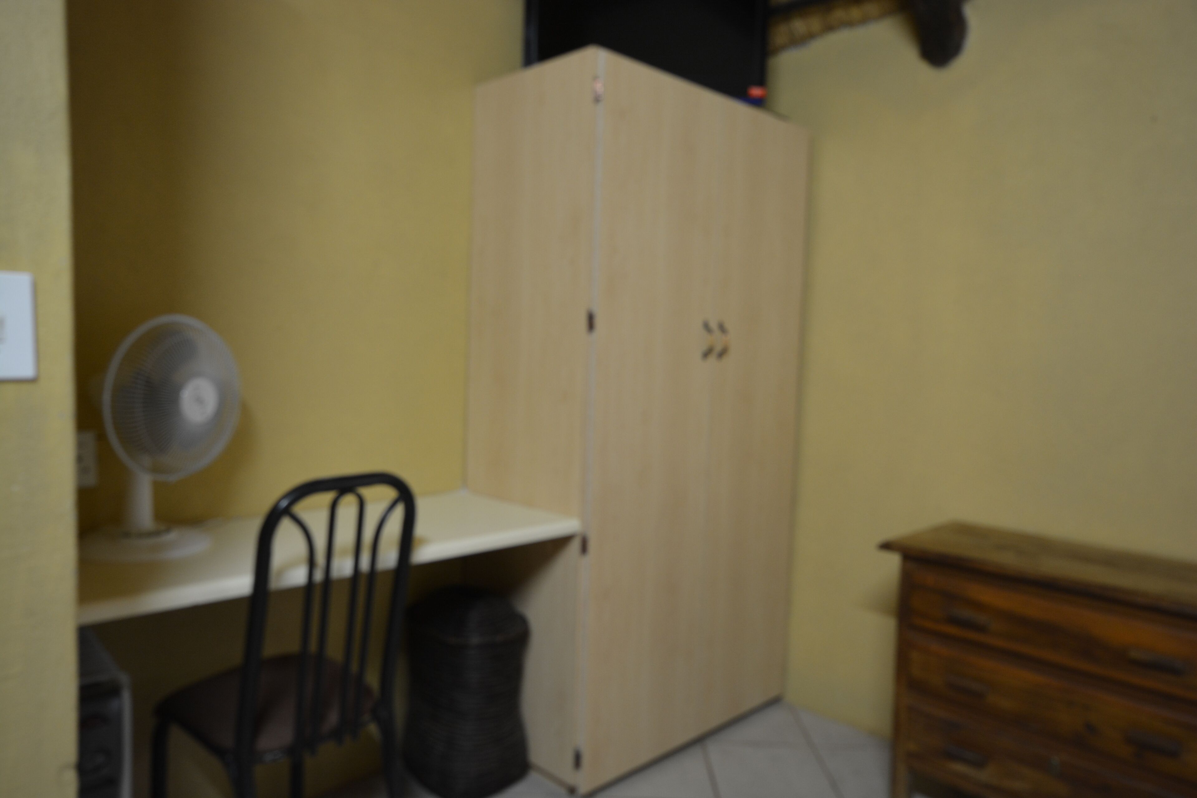 Una habitació petita amb un armari de fusta, una cadira, un escriptori amb un ventilador i un gresol.