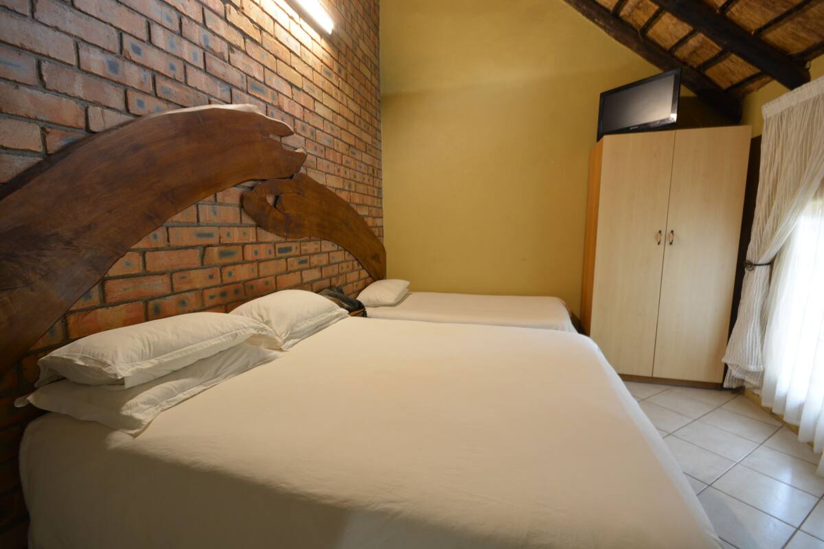 A room with a brick wall, a wooden arch, a bed with white bedding, a wardrobe, and a television on the wall.