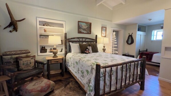 Une chambre à coucher avec un lit en bois, une table de chevet, une lampe, une chaise, une petite table et une étagère ornée d’objets décoratifs.