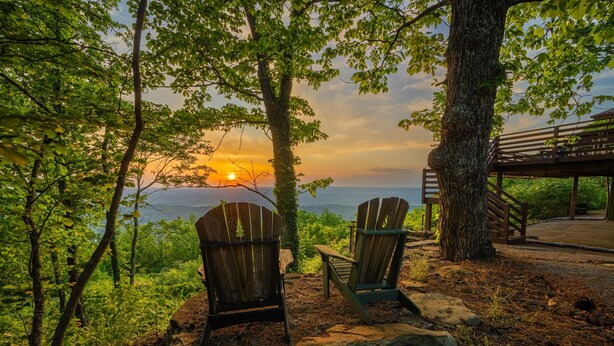 Due poltrone Adirondack rivolte verso un tramonto su un corpo d'acqua, circondate da alberi e un molo di legno.