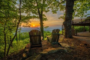 Deux fauteuils Adirondack sont placés face à un coucher de soleil sur une étendue d’eau, entourés d’arbres et d’une terrasse en bois.