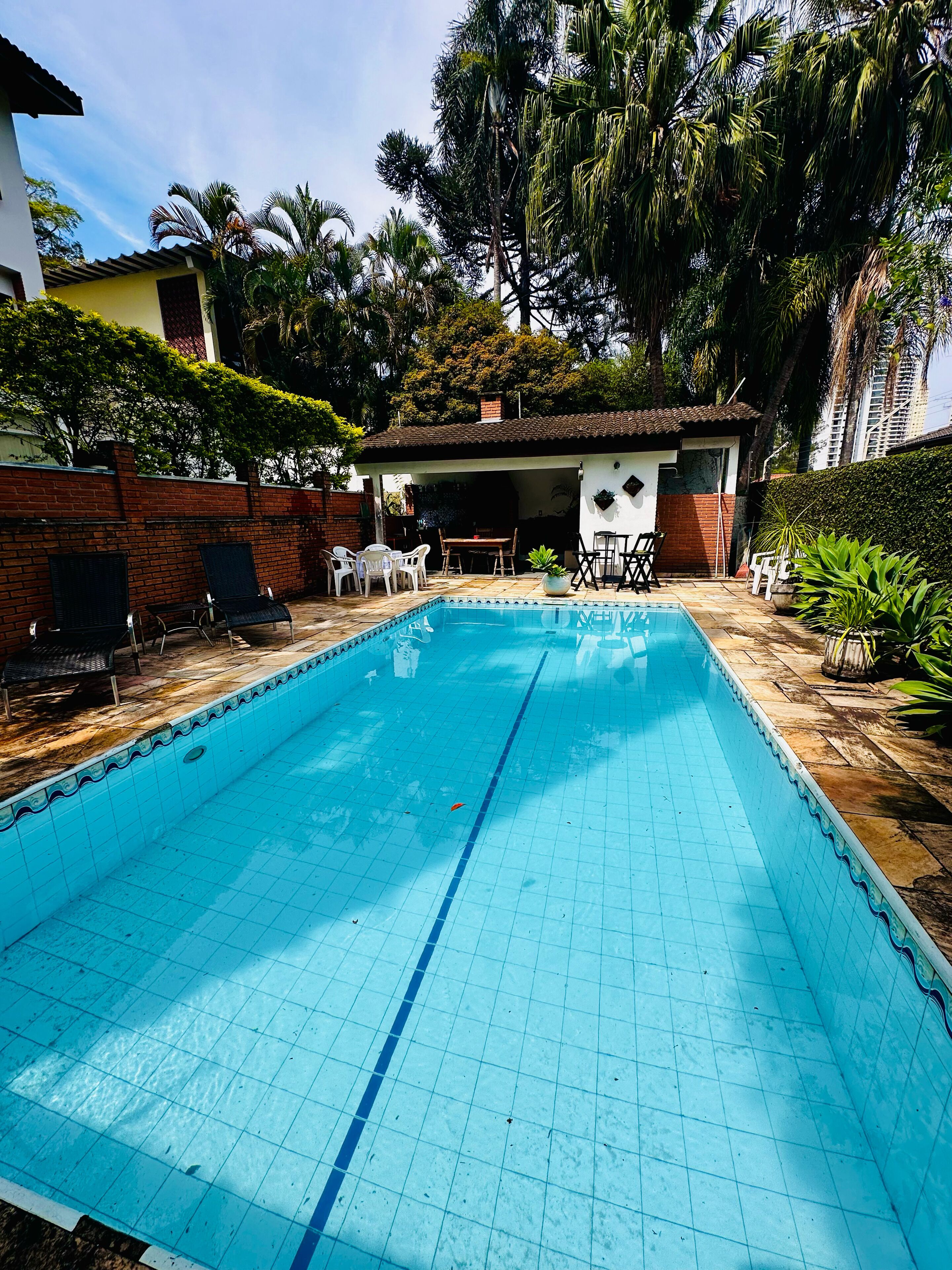 A rectangular swimming pool with tiled edges, surrounded by a brick wall and a small covered seating area with chairs and a table.