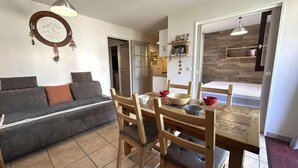 Een gezellige woonkamer met een bank, een houten eettafel en stoelen. Een keuken met kasten en een aanrecht is zichtbaar door een open doorgang.