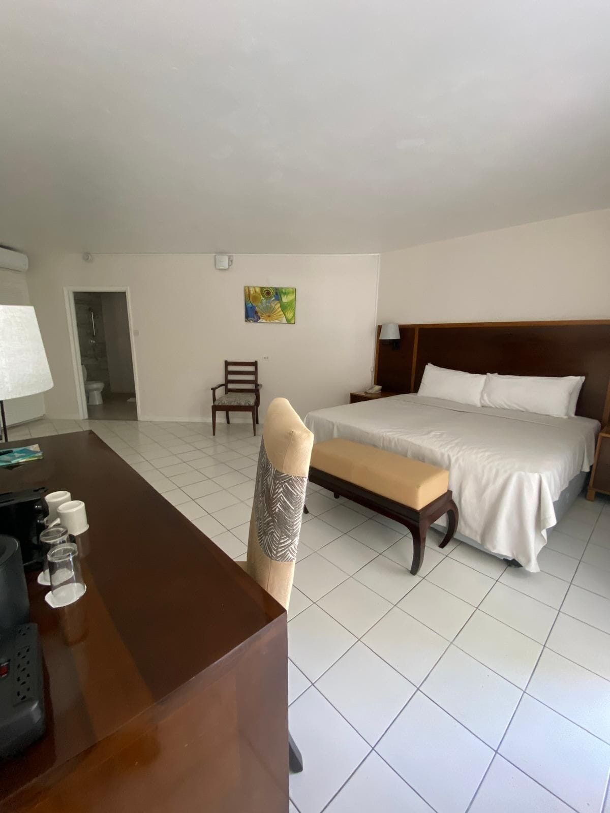 A hotel room with a bed, a desk with a coffee maker, a chair, and a painting on the wall.