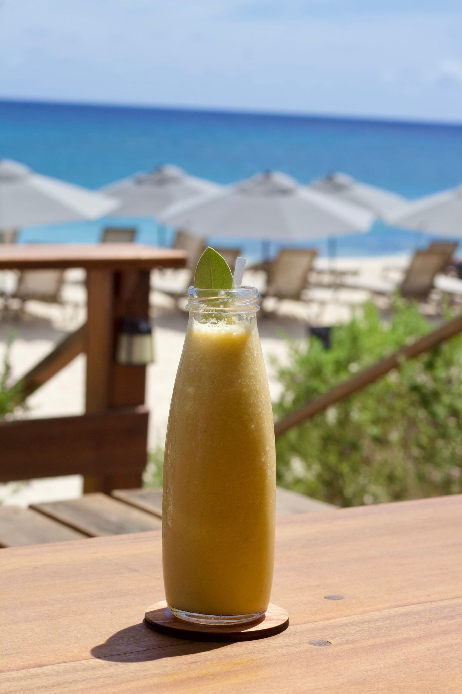 A tall glass of yellow beverage on a wooden table with a view of the beach and umbrellas in the background.