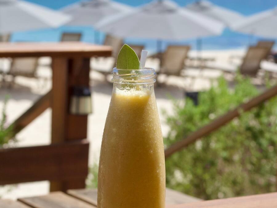 A tall glass of yellow beverage on a wooden table with a view of the beach and umbrellas in the background.