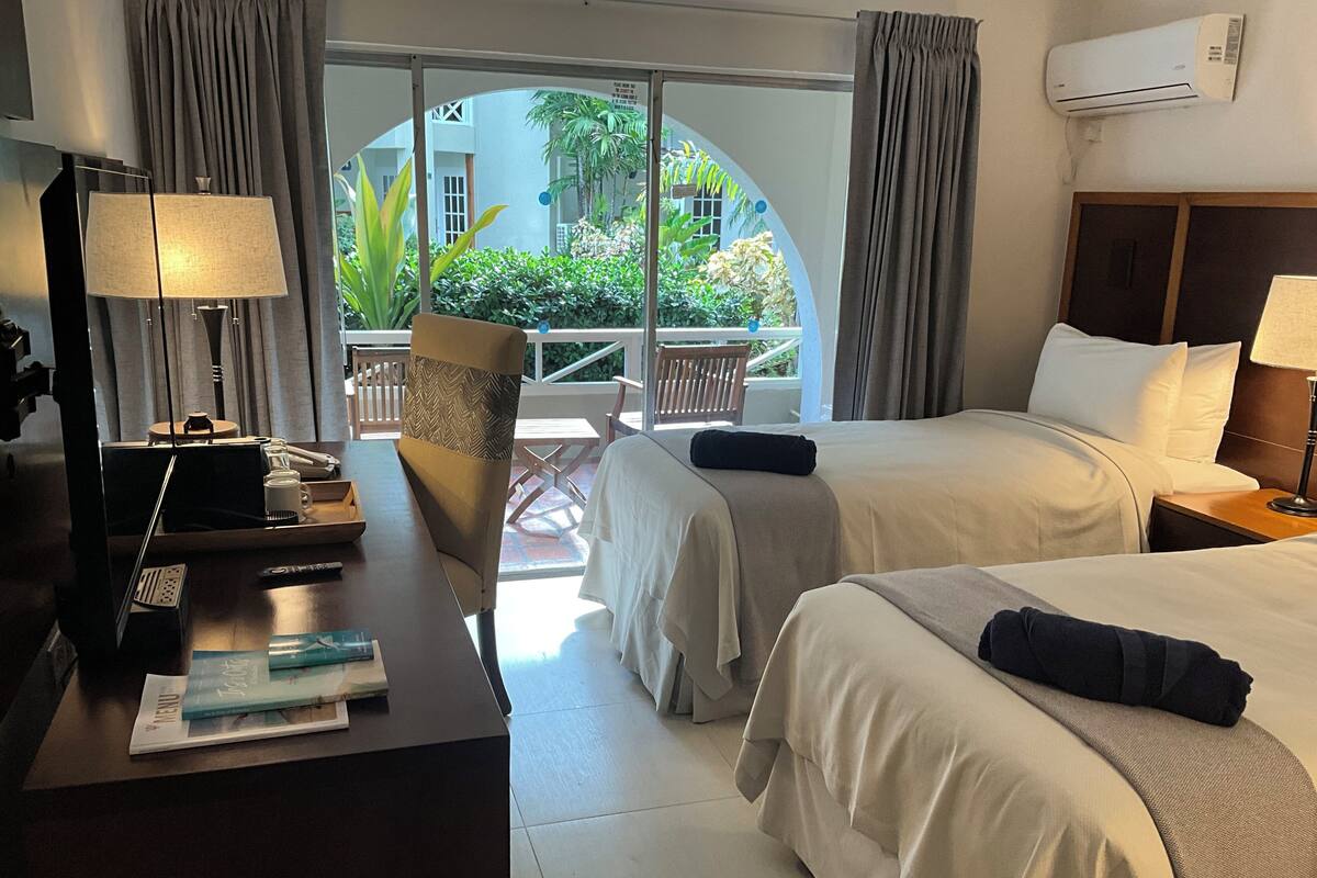 Ein Hotelzimmer mit zwei Betten, einem Schreibtisch mit Lampe, einem Sessel und Blick auf einen Innenhof mit Pflanzen und einem Gebäude.