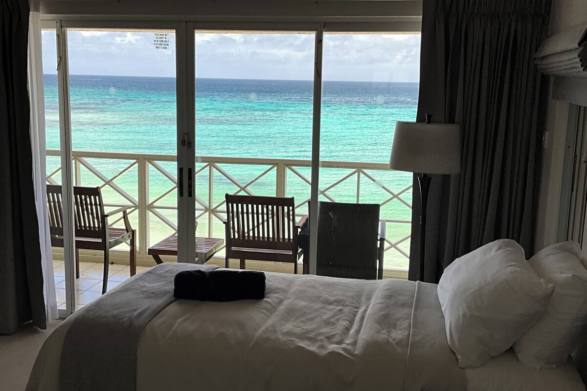 Ein Hotelzimmer mit Bett, Nachttisch, Lampe und Meerblick durch ein großes Fenster.