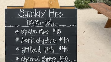 Tabla de menú de un restaurante frente a la playa, con platos como pescado a la parrilla y pollo al estilo jerk.