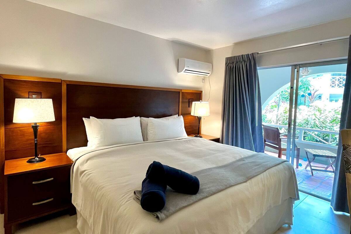 Ein Hotelzimmer mit einem großen Bett, zwei Nachttischlampen, einem hölzernen Kopfteil, einem Nachttisch und Blick ins Freie durch eine Schiebeglasdoor.