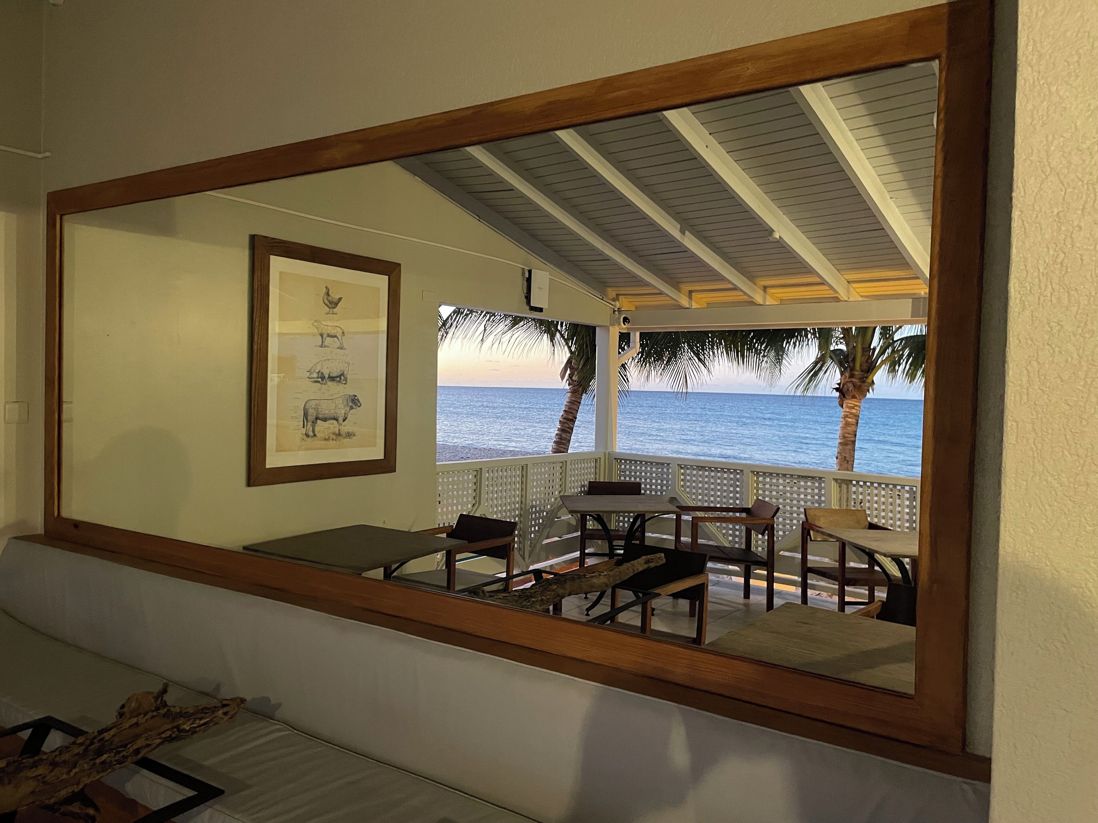A room with a large mirror reflecting a view of the ocean and palm trees. There is a framed animal illustration on the wall, a wooden bench, and a table with chairs.