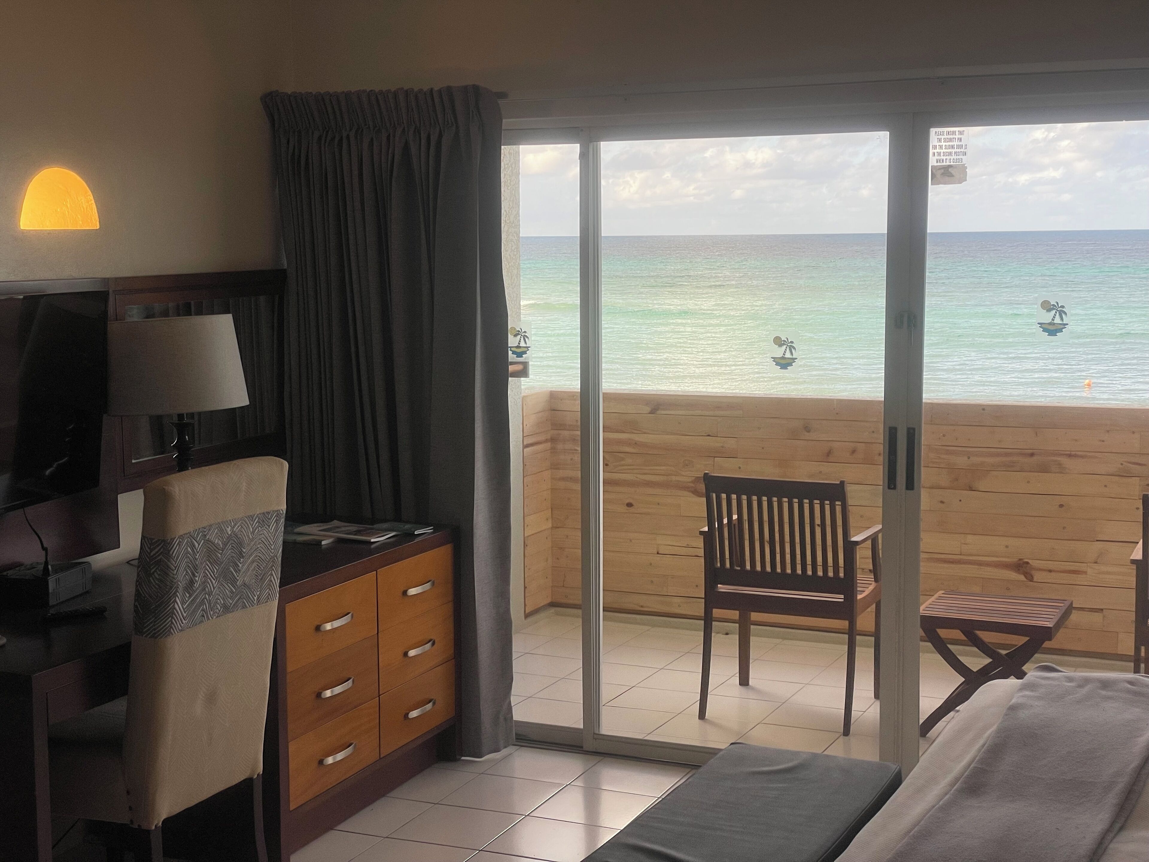 A hotel room with a bed, a desk with a lamp, a chair, and a view of the ocean through a sliding glass door.
