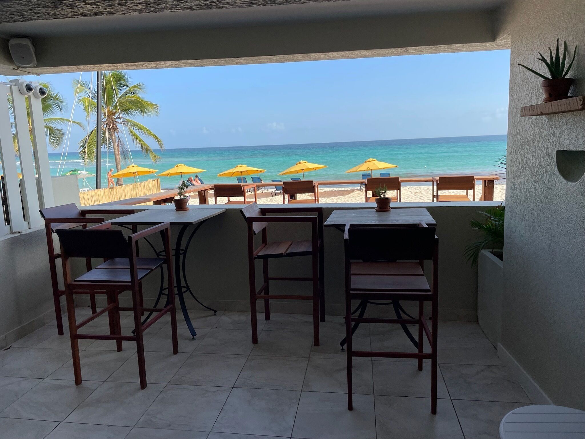 A terrace with wooden tables and chairs, overlooking a beach with yellow umbrellas and palm trees.