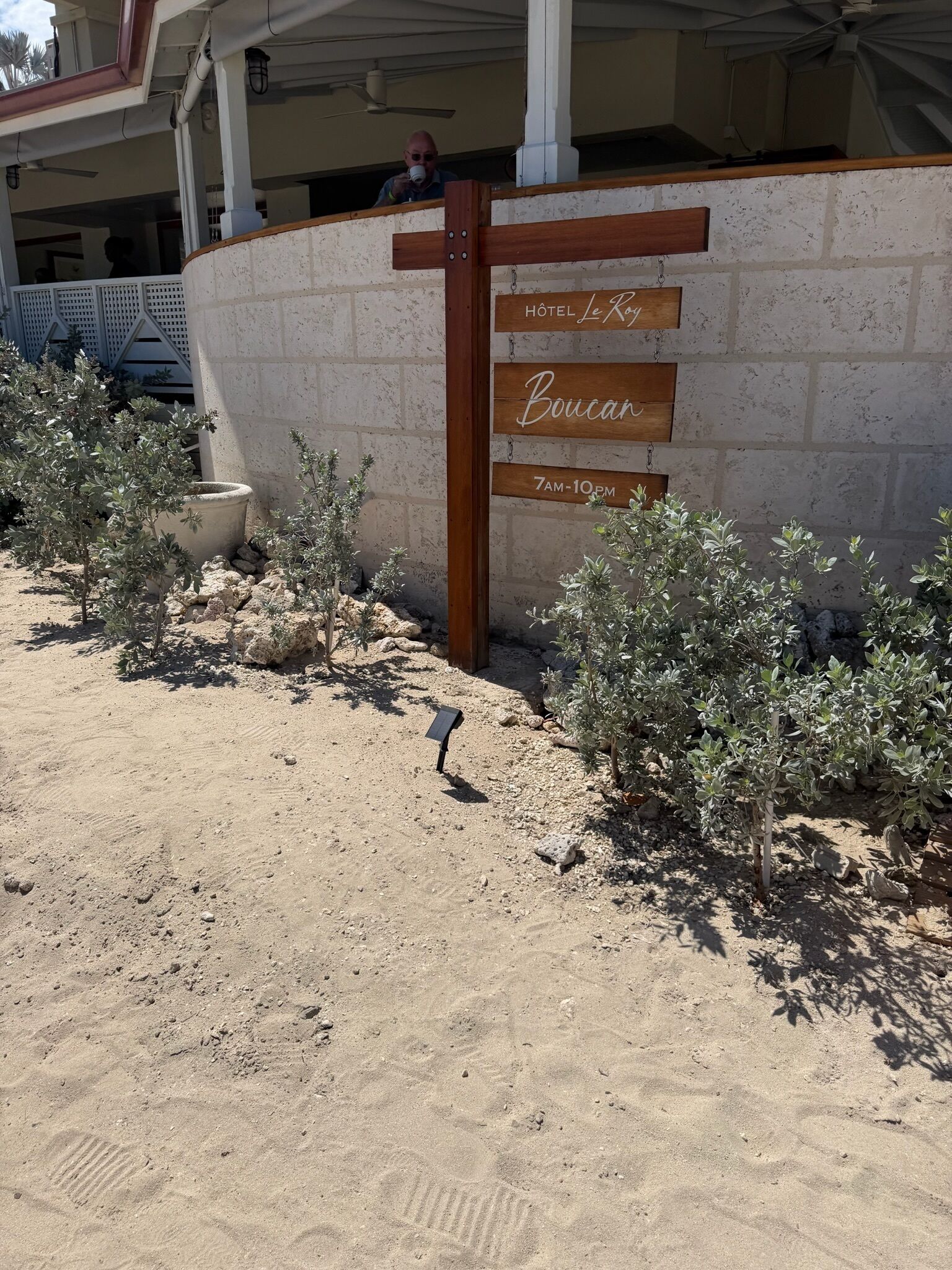 Un cartel de hotel con 'Hôtel le Roy' y 'Boucan' escritos en tablones de madera, una pared de concreto y pequeños arbustos.