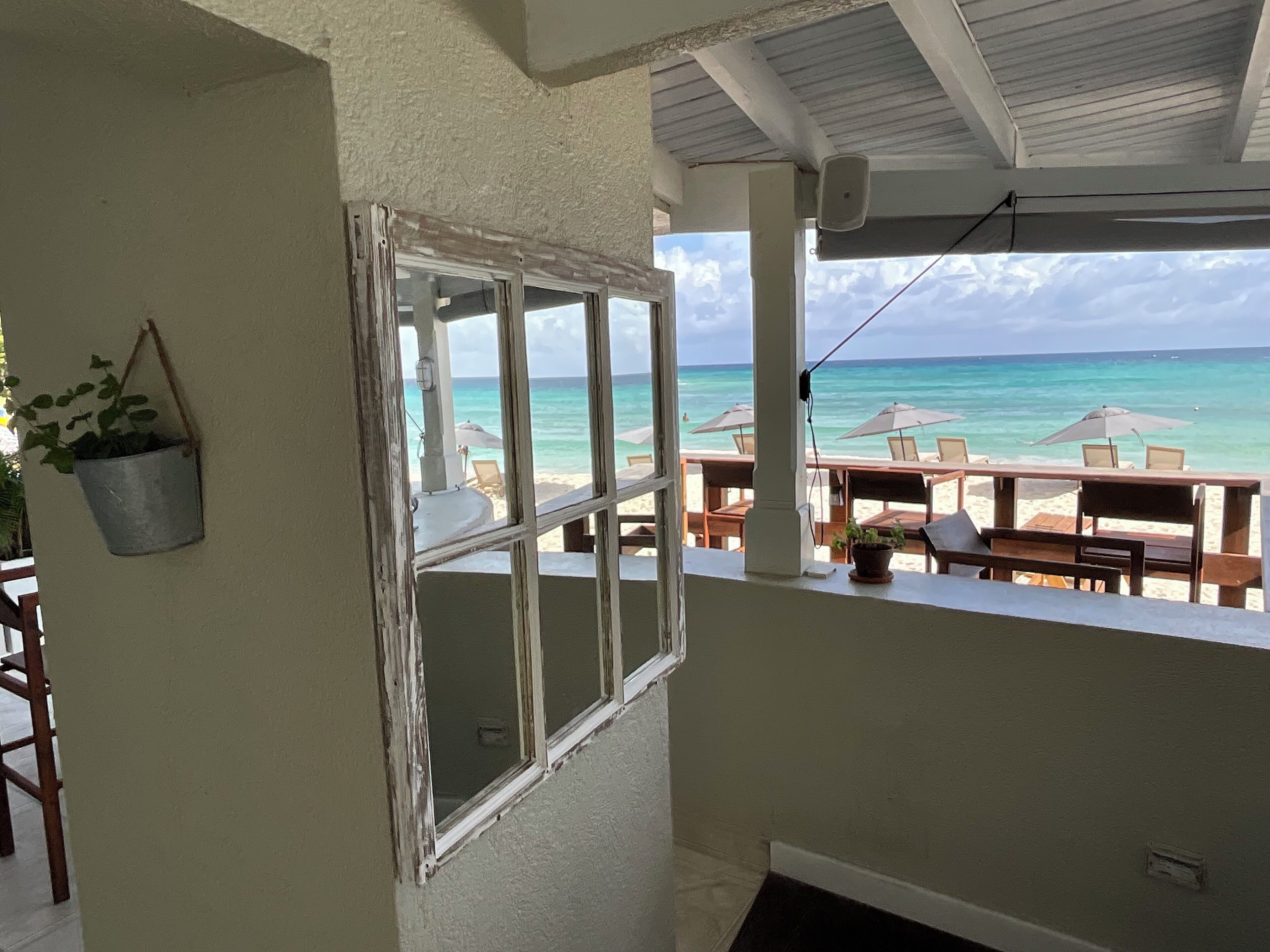 A balcony with a view of the beach and ocean. There are tables and chairs, and a hanging plant.