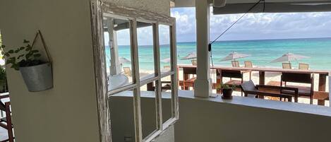 A balcony with a view of the beach and ocean. There are tables and chairs, and a hanging plant.