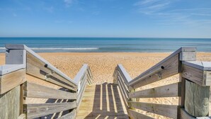 Ein hölzerner Steg führt zu einem Sandstrand, im Hintergrund das Meer.