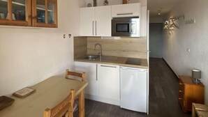 Kompakti keittiö, jossa on valkoiset kaapit, mikroaaltouuni ja pesuallas. Keittiössä on ruokapöytä tuoleineen sekä pieni puinen kaappi.