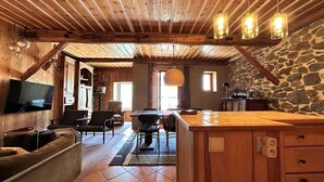 Un interior rústico con techo de madera, pared de piedra, una zona de comedor con mesa y sillas, una encimera de cocina y un televisor montado en la pared.