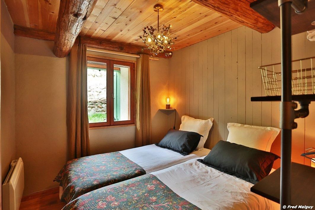 A bedroom with two beds, wooden ceiling beams, a window with curtains, a chandelier, and a bedside table with a lamp.