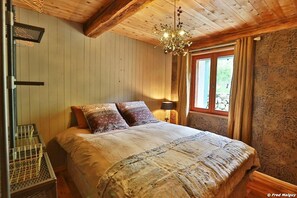 A cozy bedroom with a wooden ceiling, a large bed with patterned bedding, a chandelier, and a window with curtains.