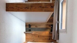 Ein Schlafzimmer im Loftsstil mit schrägverdeckter Decke, Holzbalken, einem kleinen Fenster mit Vorhängen und einer weißen Matratze.
