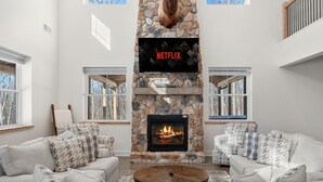 Una sala de estar con chimenea de piedra, un televisor de pantalla plana montado encima y dos sofás grises con cojines estampados.