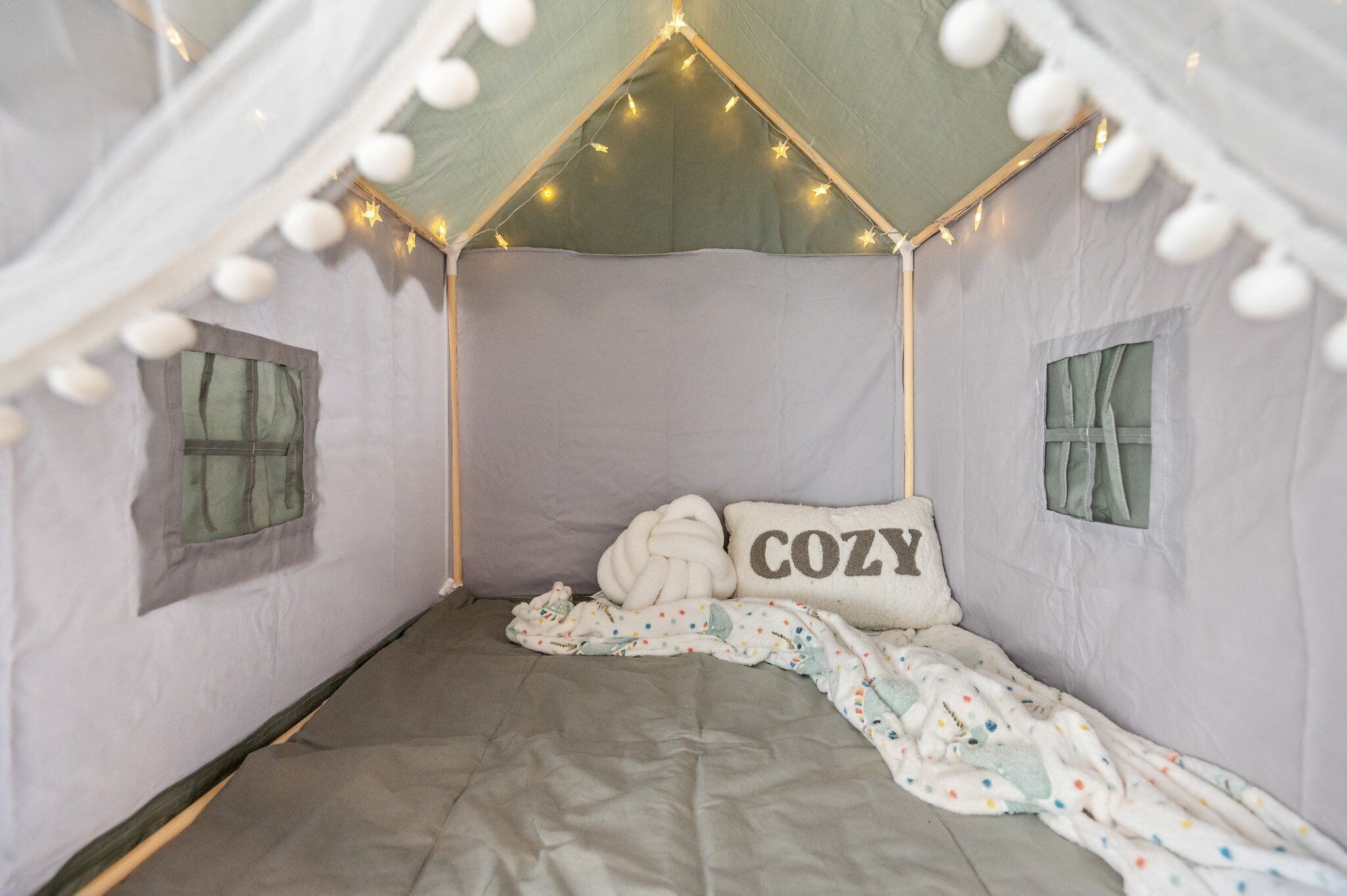 A cozy tent-like room with a bed, a pillow labeled "COZY," and string lights.