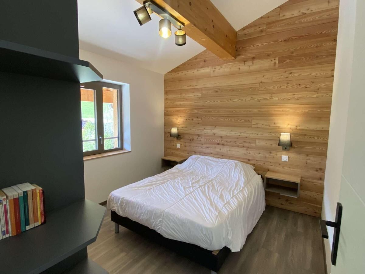 A bedroom with a wooden sloped ceiling, a bed with white bedding, two wall-mounted lamps, and a bookshelf with books.