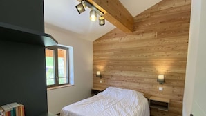 Een slaapkamer met een houten schuine daklijn, een bed met wit beddengoed, twee wandlampen en een boekenplank vol boeken.