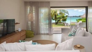 A modern living room with a television, a sofa, and a view of a tropical beach. - Unique Villa Feet in the water-pool- Grand Baie (Grand Baie)