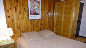 A bedroom with a wooden wall, a bed with a beige blanket, a nightstand with a lamp, and a poster of Matisse.