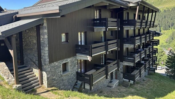 Un edificio de varios pisos con balcones, fachada de piedra y montañas al fondo.